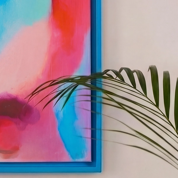 Blauwe lijst met een kleurrijke afbeelding in roze en turquoise aan een lichtbeige muur. Een groen palmblad steekt in de afbeelding.